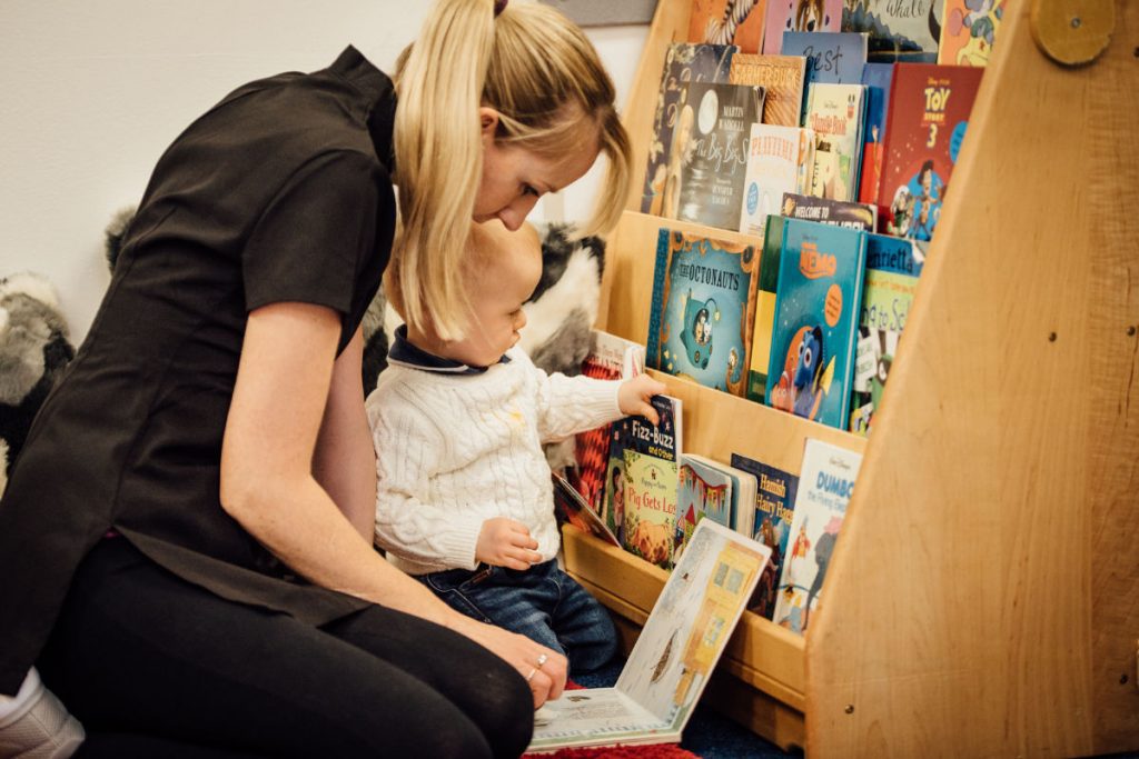 Nursery care worker sitting with baby infront of a small bookshef and reading a book