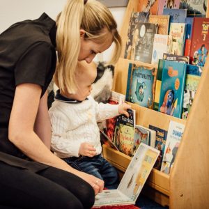 Nursery care worker sitting with baby infront of a small bookshef and reading a book